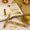 Leonardo × @twenty_sides Momento Zero Grande 2.0 Midas Fountain Pen - Fountain Pen, Jewelry, and Journal On Top Of Table | EndlessPens